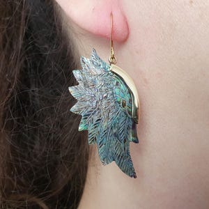 Op de afbeelding: Close-up van een oorbel met een veervormig ontwerp. De oorbel heeft een goudkleurige haak en een gedetailleerde, iriserende, blauwgroene veervorm. De oorbel wordt gedragen op het oor van een persoon.