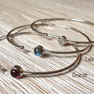 Peut inclure: Quatre bracelets en argent avec des pierres précieuses de différentes couleurs. Les bracelets sont empilés les uns sur les autres. Les pierres précieuses sont rouge, bleu, gris et blanc. Les bracelets sont étiquetés "Granat", "Labradorite", "Moonstone" et "Granat".