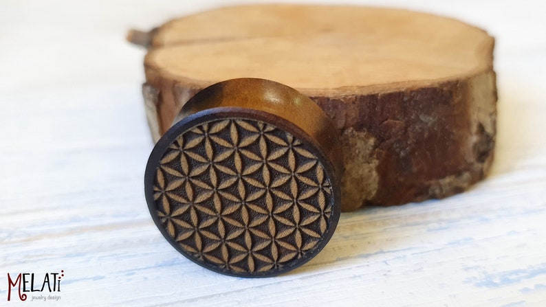 May include: A wooden ear gauge with a brown wood finish and a carved flower of life pattern.