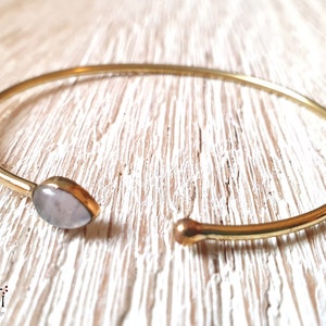 Pode incluir: Um bracelete de ouro com uma pedra oval de cor rosa claro. O bracelete é aberto e tem uma extremidade arredondada.