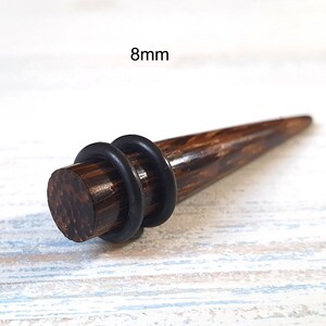 May include: A dark brown wooden ear taper with a smooth, polished finish and a pointed end. The jewelry features two black rubber O-rings positioned around the cylindrical base. The text 8mm is clearly visible above the object.