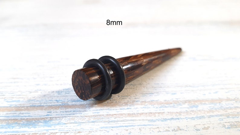 May include: A dark brown wooden ear taper with a smooth, polished finish and a pointed end. The jewelry features two black rubber O-rings positioned around the cylindrical base. The text 8mm is clearly visible above the object.