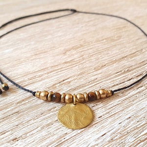 Könnte beinhalten: Eine handgefertigte Halskette mit schwarzem Band, goldfarbenen Perlen und einem strukturierten, runden Anhänger. Die Halskette enthält auch kleine, braune Perlen und wird auf einer hellen Holzoberfläche präsentiert. Schmuck von Melati Jewelry Design.