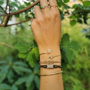Könnte beinhalten: Eine Hand trägt ein goldenes Armband mit einem Spiraldesign und einem kleinen Pfeil-Anhänger. Das Armband ist mit zwei weiteren Armbändern übereinander gelegt, eines mit einem gewebten Design in Braun und Beige und einem quadratischen Stein, das andere ein schlichtes schwarzes geflochtenes Armband. Die Hand trägt auch drei goldene Ringe.