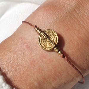 May include: A brown string bracelet with a gold circular charm featuring a concentric circle design.