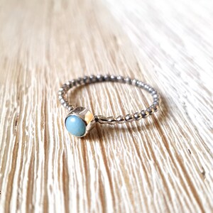 May include: A silver ring with a light blue gemstone. The band has a textured, beaded design. The gemstone is round and set in a square bezel. The ring is displayed on a light wood surface.