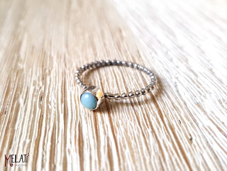 May include: A silver ring with a light blue gemstone. The band has a textured, beaded design. The gemstone is round and set in a square bezel. The ring is displayed on a light wood surface.