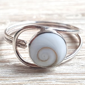 Puede incluir: Un anillo de plata con una incrustación de concha blanca. El anillo tiene un diseño simple y moderno con una sola concha redonda engastada en una banda de plata.