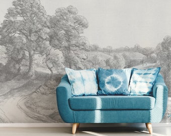 Op de afbeelding: Een blauwe bank met drie tie-dye kussens staat voor een muur met een zwart-witte tekening van een landschap. Een blauwe schommelstoel staat op de voorgrond.