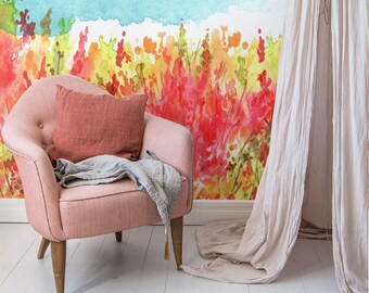 Op de afbeelding: Een roze fauteuil met een rood kussen en een grijze deken staat voor een aquarelmuurschildering die een bloemenveld en een blauwe lucht voorstelt.