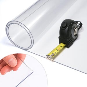 Könnte beinhalten: Gerollte, transparente Kunststofffolie mit einem schwarzen Maßband. Das Maßband zeigt Zoll an. Eine Hand hält eine Ecke einer flachen Folie aus demselben transparenten Kunststoff.