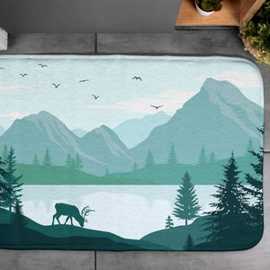 Op de afbeelding: Een blauwe en groene badmat met een berglandschap en een hert-silhouet. De mat heeft een meer met een reflectie van de bergen.