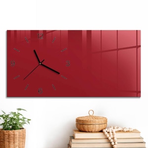 May include: A rectangular wall clock with a deep red, glossy surface. The clock face features black hour and minute hands, with black numerical markers for 3, 6, 9, and 12, and simple black tick marks for the remaining hour positions.