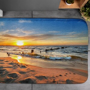 May include: A bath mat depicting a sunset over the ocean. The mat shows a sandy beach with footprints and a wave breaking on the shore. The sky is a vibrant orange and pink with clouds.