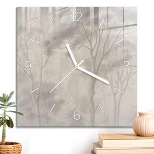 May include: A square wall clock featuring a muted beige and gray forest scene with bare trees. The clock face displays white hour markers, white numerals at the 12, 3, 6, and 9 positions, and two white clock hands centered on the printed design.