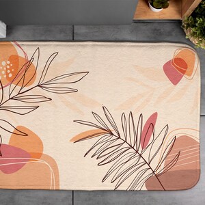 Op de afbeelding: Rechthoekige badmat met een beige achtergrond en een abstract ontwerp. Het ontwerp bevat bruine lijntekeningen van bladeren en abstracte vormen in tinten oranje en roze. De mat is ontworpen voor gebruik in de badkamer.