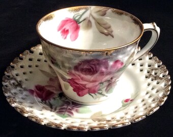 Puede incluir: Un juego de taza y platillo de té blanco con un delicado diseño de encaje y ribete dorado. La taza y el platillo están decorados con rosas rosas y moradas.