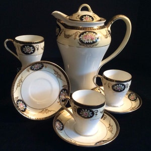 Op de afbeelding: Een vintage theeservies met een crèmekleurige basis en gouden accenten. De set bevat een theepot, vier kopjes en vier schoteltjes. De kopjes en schoteltjes hebben een bloemmotief met roze en zwarte accenten.