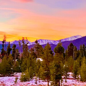 Puede incluir: Un paisaje pintoresco con una puesta de sol sobre montañas nevadas y un bosque de árboles de hoja perenne. El cielo transita del naranja al rosa, creando un telón de fondo colorido para la escena natural.