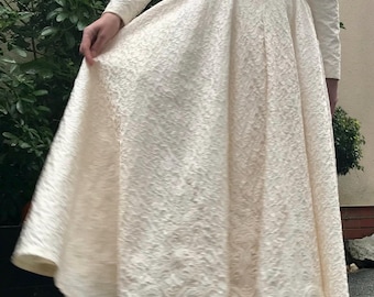 Puede incluir: Un vestido blanco de manga larga con escote en V y un corpiño ajustado. La falda es amplia y fluida, y el vestido tiene un cinturón en la cintura. El vestido está hecho de una tela texturizada.