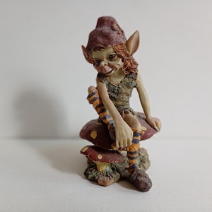 May include: A small figurine of a creature with pointed ears, reddish hair, and a dark cap, wearing a textured green top and striped orange and blue leggings. The creature is seated on a pair of brown mushrooms with yellow spots on a base.