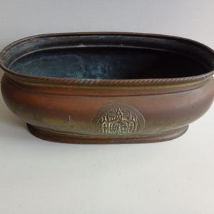 May include: An oval-shaped copper planter with a dark, weathered patina and a rope-style rim. The exterior features a centred, embossed medallion depicting a building with multiple spires. The interior is dark and shows signs of oxidation.