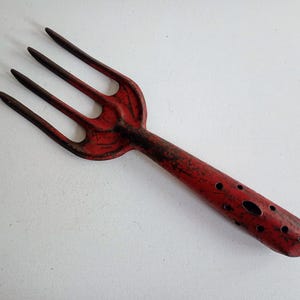 May include: A vintage red garden fork with four prongs and a long handle. The handle features several circular holes. The metal tool shows signs of wear, with a distressed finish. A gardening tool.