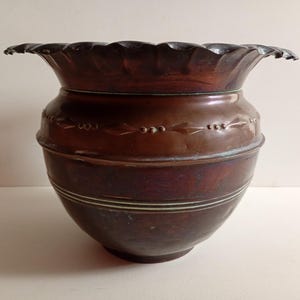 May include: A dark copper-toned metal planter with a wide, scalloped rim. The body features a horizontal band of embossed diamond and dot patterns, with two thin, parallel engraved lines encircling the lower section of the rounded vessel.