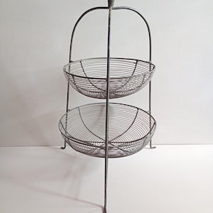 May include: A two-tiered wire basket stand featuring a dark grey metal finish. The structure consists of two circular wire mesh bowls stacked vertically, supported by a central metal frame with a handle at the top and three thin metal legs.