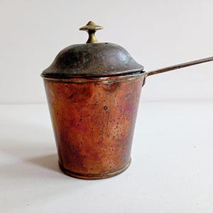 May include: An antique copper pot with a lid and a long handle. The pot has a cylindrical body and a slightly domed lid with a small brass knob. The copper has a rich, aged patina, with variations in colour from reddish-brown to darker hues.