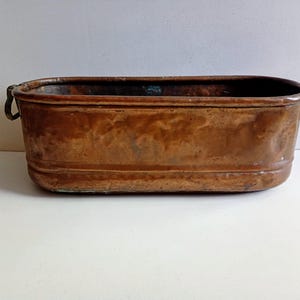 May include: A rectangular copper planter with a weathered patina. The planter has rounded corners, a slightly raised base, and small, curved handles on each end. The copper has a rich, warm brown colour with darker areas, suggesting age and use.