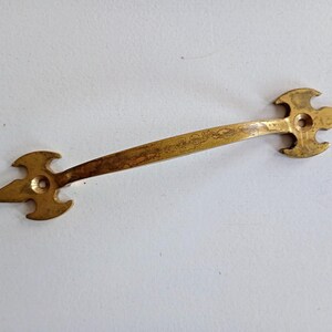 May include: A vintage brass drawer pull with an ornate design. The pull features a long, slender handle with decorative arrow-shaped ends. The brass has a warm, aged patina, suggesting its antique nature. The hardware is likely for a cabinet or drawer.