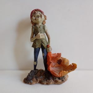 May include: A small figurine of a smiling character wearing a red cap, a textured green vest, and dark blue trousers with patches. The figure stands on a brown base and holds a large, orange, leaf-shaped object in one hand while resting the other hand on their chest.