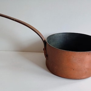 May include: A round copper saucepan with a long, curved metal handle attached by rivets. The exterior features a warm, metallic copper finish with visible signs of wear, while the interior has a dark, matte grey lining and a smooth rim.