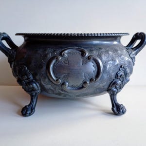 May include: An antique silver-toned bowl with intricate floral and scrollwork. It features two curved handles and stands on three claw feet. The rim has a textured, beaded design. A central cartouche is visible on the front.