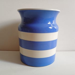 May include: A cylindrical ceramic vase featuring a glossy finish with alternating horizontal stripes in bright blue and white. The vase has a slightly flared rim at the top and a smooth, rounded body, showcasing a clean design with no additional patterns or textures.