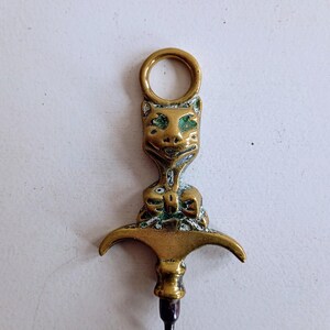 May include: A vintage brass corkscrew featuring a cat-shaped handle. The cat's face is detailed with etched features and a ring at the top. The corkscrew has a bow tie design and a dark metal screw.