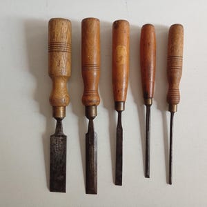 May include: A set of five wood chisels arranged in a row, each featuring a light brown wooden handle with a cylindrical shape and a metal ferrule connecting to a dark, weathered steel blade of varying widths, from wide to narrow.