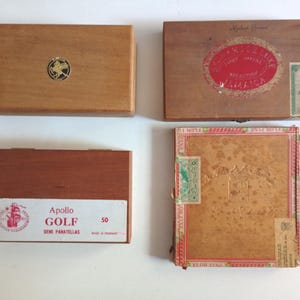 May include: Four rectangular wooden cigar boxes arranged in a grid. One box features a gold circular emblem, another has a red oval label reading Jamaica, a third is labelled Apollo Golf Demi Panatellas 50, and the fourth has a tan textured finish.