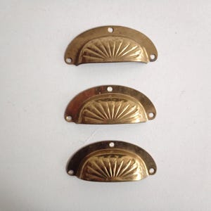 May include: Three semi-circular brass drawer pulls arranged vertically. Each pull features a central recessed area with a radiating sunburst pattern and three small circular screw holes located at the top and sides for mounting purposes.