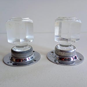 May include: A pair of clear, faceted glass door knobs with a square top and a chrome base. They are mounted on round, silver-coloured metal plates. The background is a plain, off-white surface.