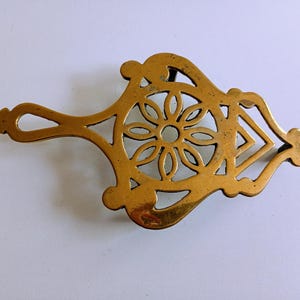 May include: A gold-coloured brass trivet with a floral centre and a handle. The trivet has an openwork design, allowing heat to dissipate. It is likely used to protect surfaces from hot cookware. The trivet measures approximately 20 cm in length.