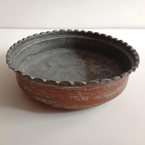 May include: A round, shallow copper bowl with a weathered, oxidised exterior and a scalloped, crimped rim. The interior surface is a dull, tarnished grey metal, showing signs of age and wear across the bottom and the curved side walls.