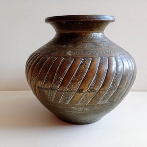 May include: A vintage bronze-coloured pot with a wide, rounded body and a narrow neck. The pot features intricate etched designs, including vertical, textured stripes and decorative bands around the neck. The aged metal has a warm, antique appearance.