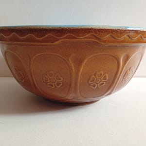 May include: A brown ceramic mixing bowl with a glossy finish, featuring a scalloped rim and embossed floral patterns around the exterior. The interior of the bowl is glazed in a bright turquoise blue, contrasting with the warm brown exterior.