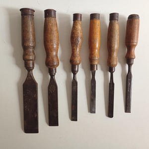 May include: A set of six wood chisels arranged in a row, each featuring a dark metal blade and a weathered wooden handle with a metal ferrule at the top. The tools vary in width and length, displaying signs of age and use on the metal and wood surfaces.