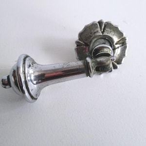 May include: A silver-toned metal drawer pull featuring a long, tapered handle with a rounded end and a circular, scalloped backplate. The hardware has a polished, reflective finish with visible signs of wear and minor dark spots on the surface.