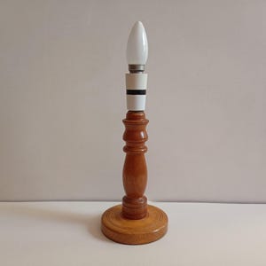 May include: A wooden table lamp with a light bulb. The lamp has a turned wood base and a round base. The light bulb is white and has a pointed tip. The lamp is made of light brown wood.