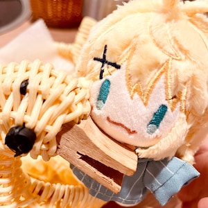 Op de afbeelding: Een pluche pop met geel haar en geborduurde gezichtskenmerken, gekleed in een blauw gestreept shirt, ligt in een gevlochten mand. De pop houdt een klein houten voorwerp vast. De mand en de pop creëren een charmante, handgemaakte esthetiek.