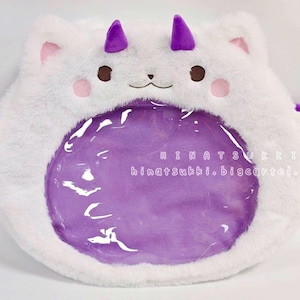 May include: A white plush toy shaped like a cat with purple wings and a horn. The toy has a clear plastic window on the front. The text "HINATSUKKI hinatsukki.bigcartel.com" is printed on the toy.